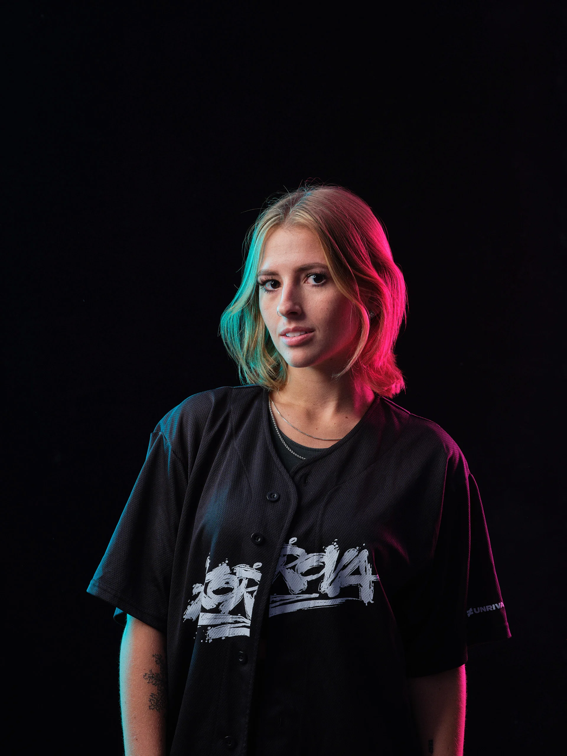 Frontal character portrait of a woman in a black graphic jersey, illuminated by teal and magenta volumetric rim lighting.