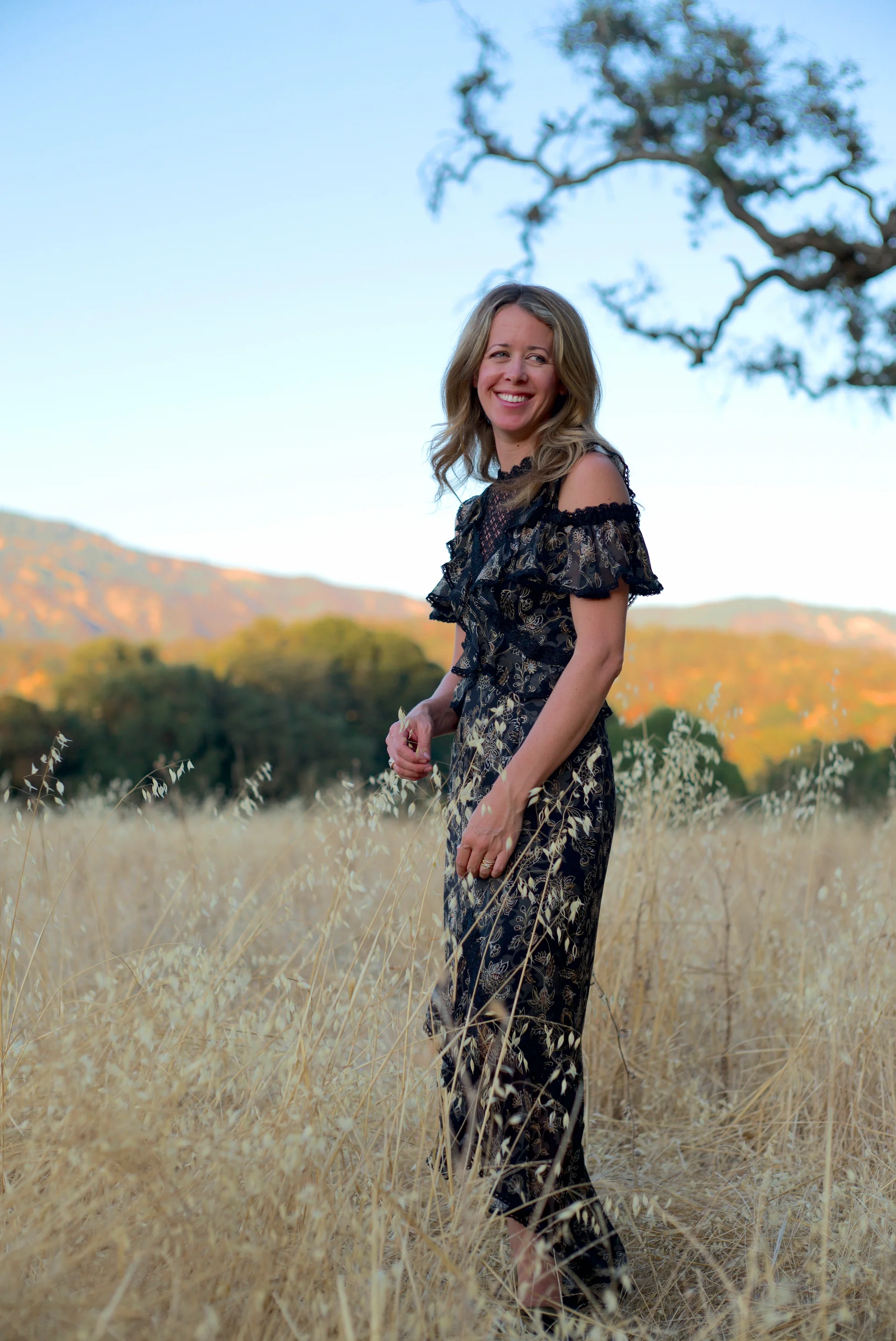 Elegant outdoor portrait by San Francisco photographer; a female winemaker in a floral dress standing in a golden Napa Valley field at sunset, captured with Leica natural light for a soft, timeless aesthetic.