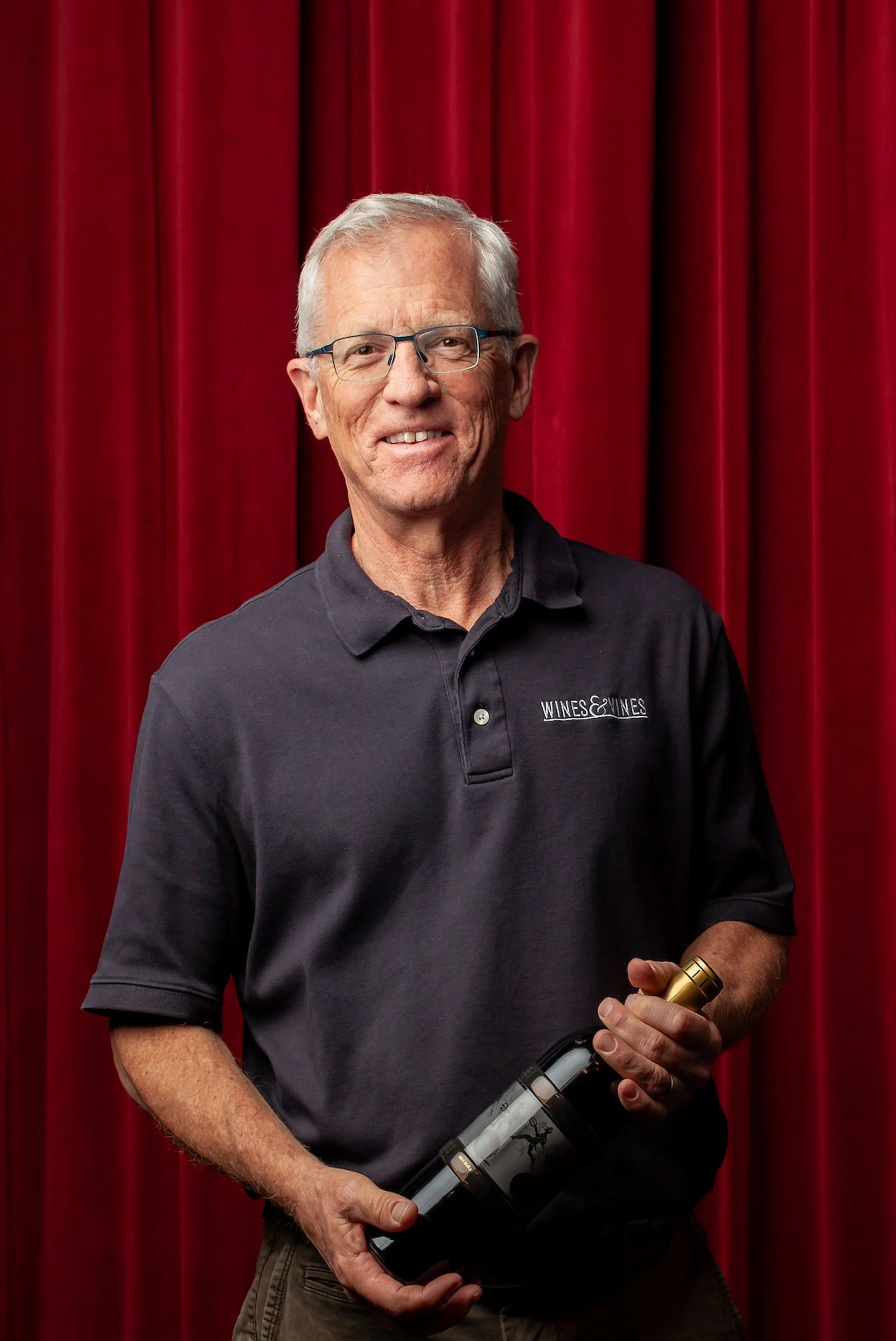 Professional event portrait by San Francisco photographer; wine label contest winner holding a trophy bottle against a red velvet curtain, captured with clean, institutional lighting.