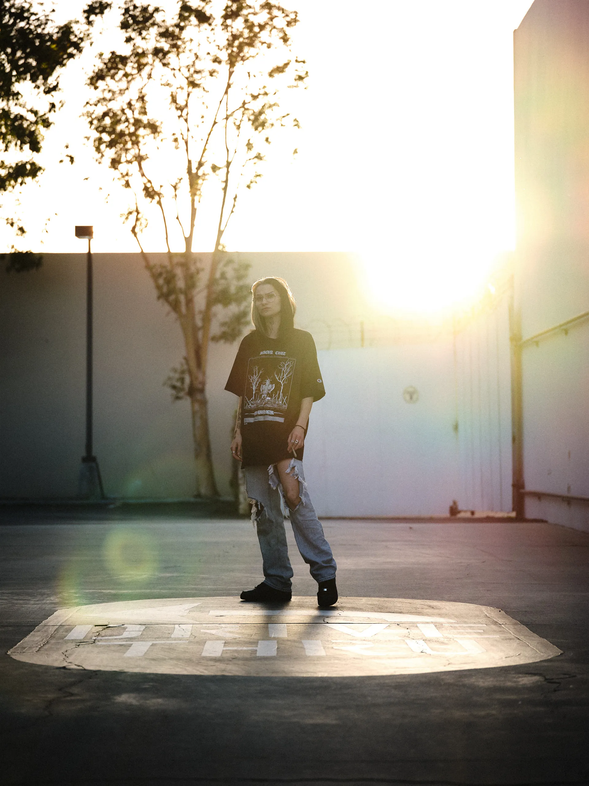 Full-body lifestyle portrait by San Francisco photographer; woman in streetwear standing in a 'Drive Thru' circle backlit by a powerful sunset, captured on GFX with cinematic lens flare.